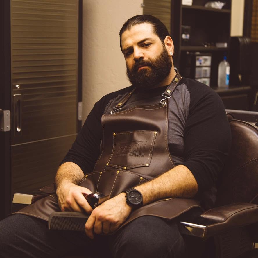 Brennan in a leather apron, sitting in his barber chair with clippers in hand